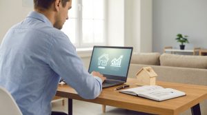 Homme recherchant un logement sur un ordinateur portable, avec des icônes de maison et de graphique, un modèle de maison et un carnet sur le bureau.